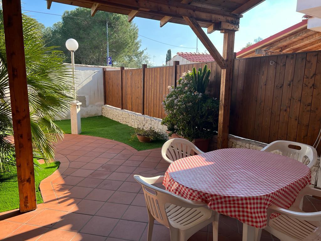 An outdoor veranda features a round table covered with a red and white checkered tablecloth, surrounded by white plastic chairs. Lush green plants and grass add a natural touch, while wooden beams provide a shaded area. A pathway leads to a parking space in the background.