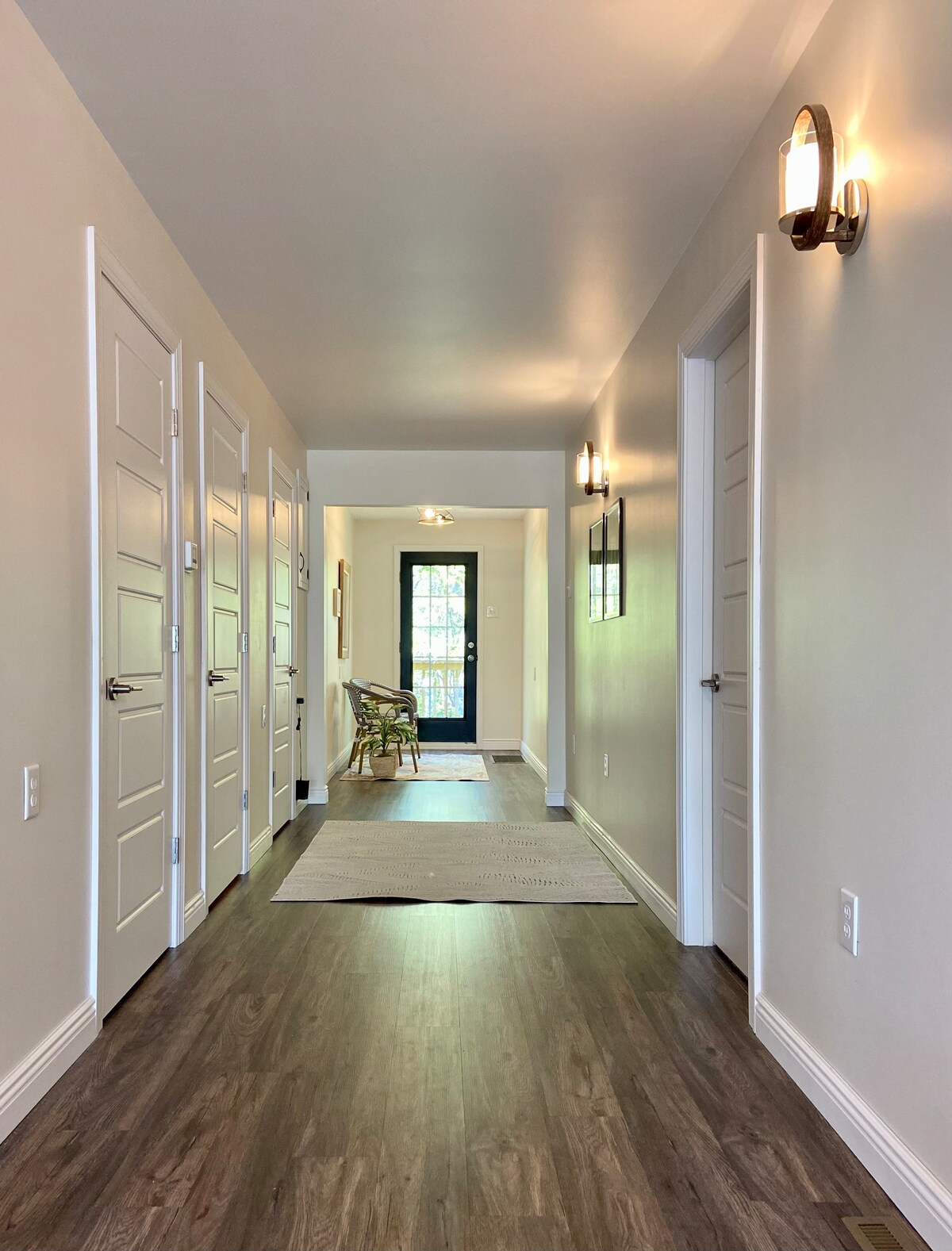 A well-lit hallway features multiple doors on either side, leading to rooms. A door at the end opens to an outdoor area. Soft lighting fixtures adorn the walls, and a subtle area rug adds warmth to the floor, which is finished in rich wood tones.