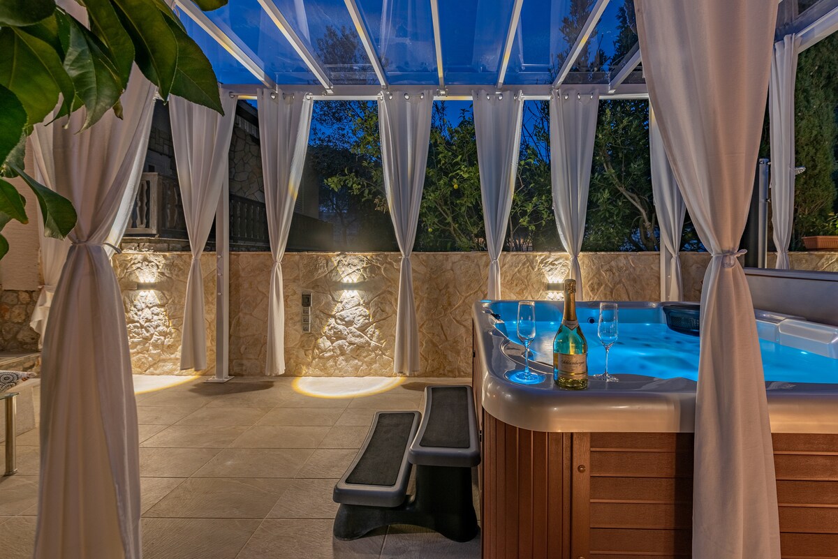An indoor relaxation area is presented, featuring a hot tub under a glass canopy. Soft lighting highlights the stone walls and potted plants, creating a serene ambiance. Two steps lead up to the hot tub, accompanied by a bottle of wine, enhancing the inviting setting.