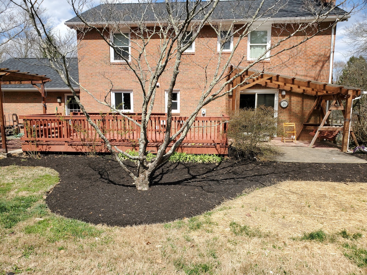 The exterior shows a brick home with a wooden deck and patio area, featuring a covered swing. Surrounding greenery includes mulch beds and a bare tree, while the lawn is well-maintained with patches of grass.