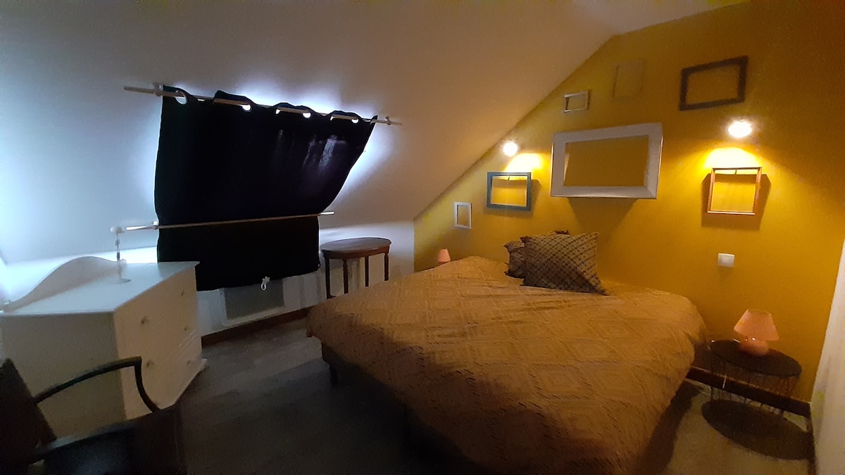 A cozy bedroom features a spacious bed adorned with a patterned blanket. A bedside table with a lamp is positioned next to the bed. Two wall-mounted lights provide soft illumination. The walls are painted yellow, and framed decorations add visual interest.