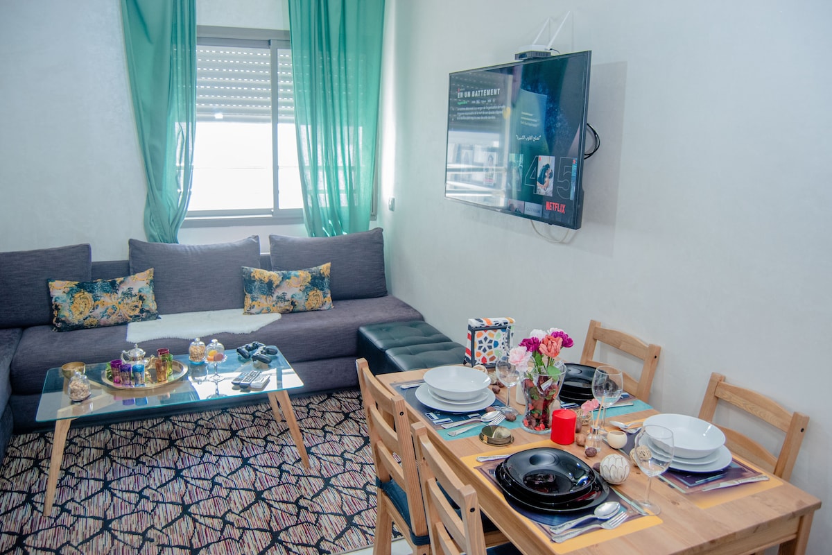 A comfortable living space features a large sofa adorned with decorative cushions and a glass coffee table displaying various items. A dining area includes a wooden table set with plates, glasses, and a vibrant centerpiece of flowers, while a wall-mounted TV adds visual interest.