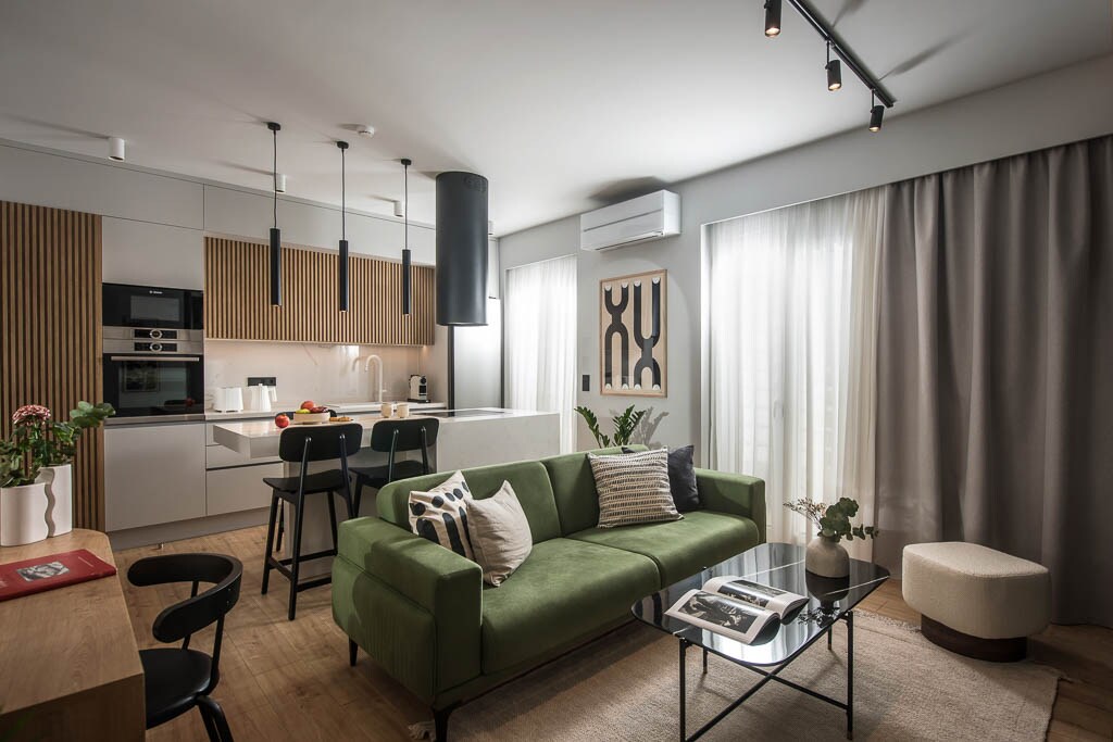 A modern living area is highlighted by a green sofa, complemented by a neutral-colored rug. Natural light is received through two tall windows, which are dressed in light curtains. A dining table is nearby, and a sleek kitchen with contemporary fixtures is visible.