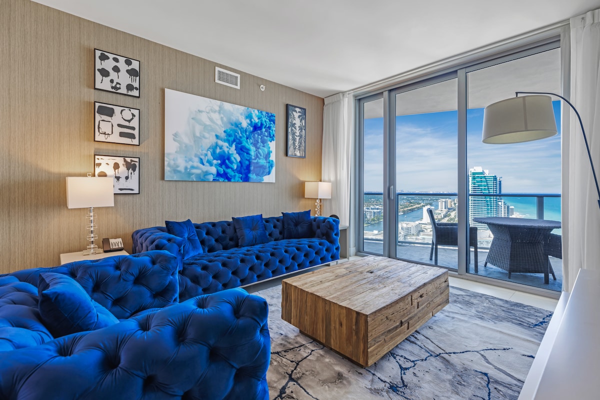 A bright and inviting living area features blue tufted sofas with a natural wood coffee table at the center. Abstract wall art complements the decor, while large glass doors provide sweeping views of the ocean and city beyond, enhancing the overall ambiance.