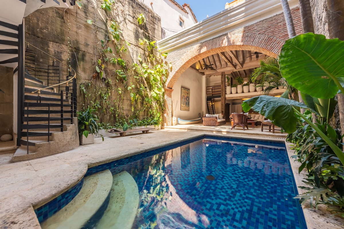An inviting pool area is surrounded by lush greenery and features tranquil blue tiles. Arched brickwork highlights the space, while a wooden deck provides relaxation zones. Natural light brightens the scene, emphasizing the blend of tropical plants and colonial design elements.