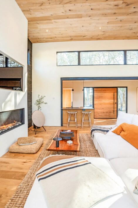 A spacious living area is defined by warm wood tones and large windows, allowing natural light to flood the room. A modern fireplace is centrally located, while a wooden coffee table sits atop a woven rug. Soft seating is arranged for comfort.