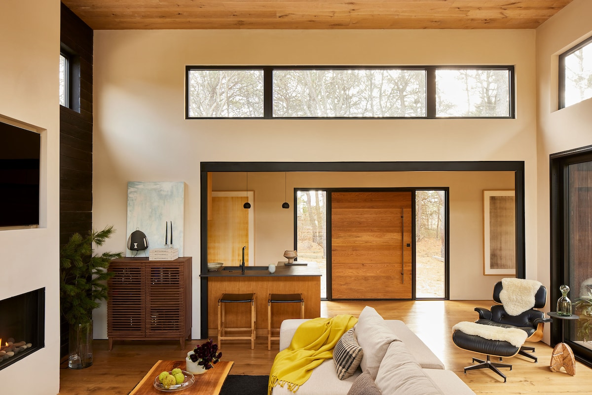 A modern living space showcases a comfortable sofa partially draped with a cozy throw. Large windows fill the area with natural light, and a wooden door adds warmth. An inviting dining area is visible, featuring minimalist furniture and subtle decorative elements.