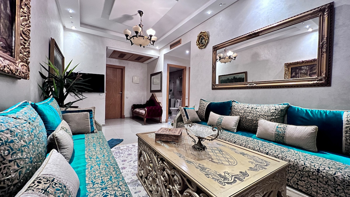 A spacious living area features elegant seating in shades of turquoise and gold. A decorative coffee table with intricate designs sits in the center, complemented by a large mirror and tasteful wall art. Soft lighting enhances the inviting atmosphere, creating a comfortable gathering space.