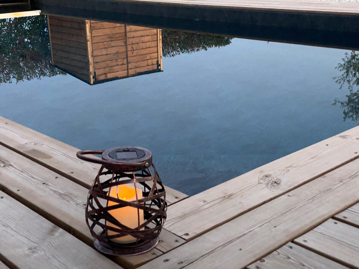 A woven lantern with a candle sits on the wooden deck beside a tranquil pool. The water reflects the surrounding greenery and warm wooden structures, creating a serene environment. Soft natural light enhances the peaceful atmosphere.