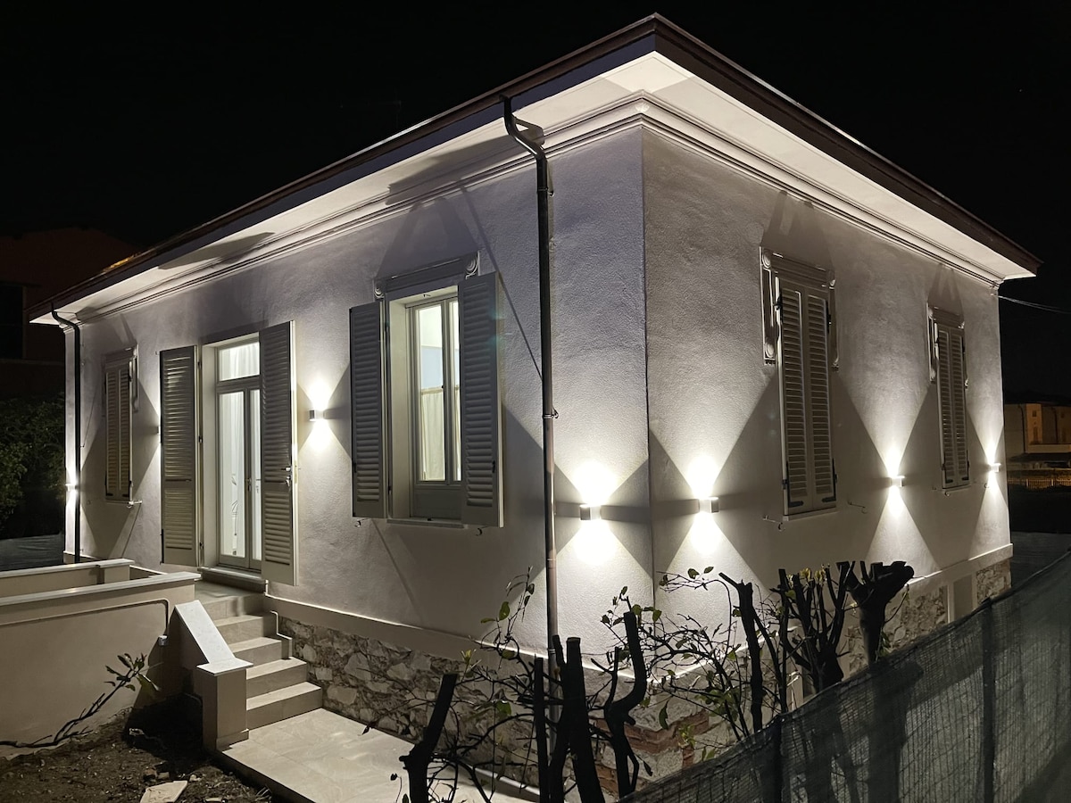 The exterior of the house is showcased at night, illuminated by strategically placed lights highlighting its elegant architecture. The facade features light-colored walls and numerous shuttered windows. A small staircase leads to the front entrance, flanked by stone details and surrounding greenery.