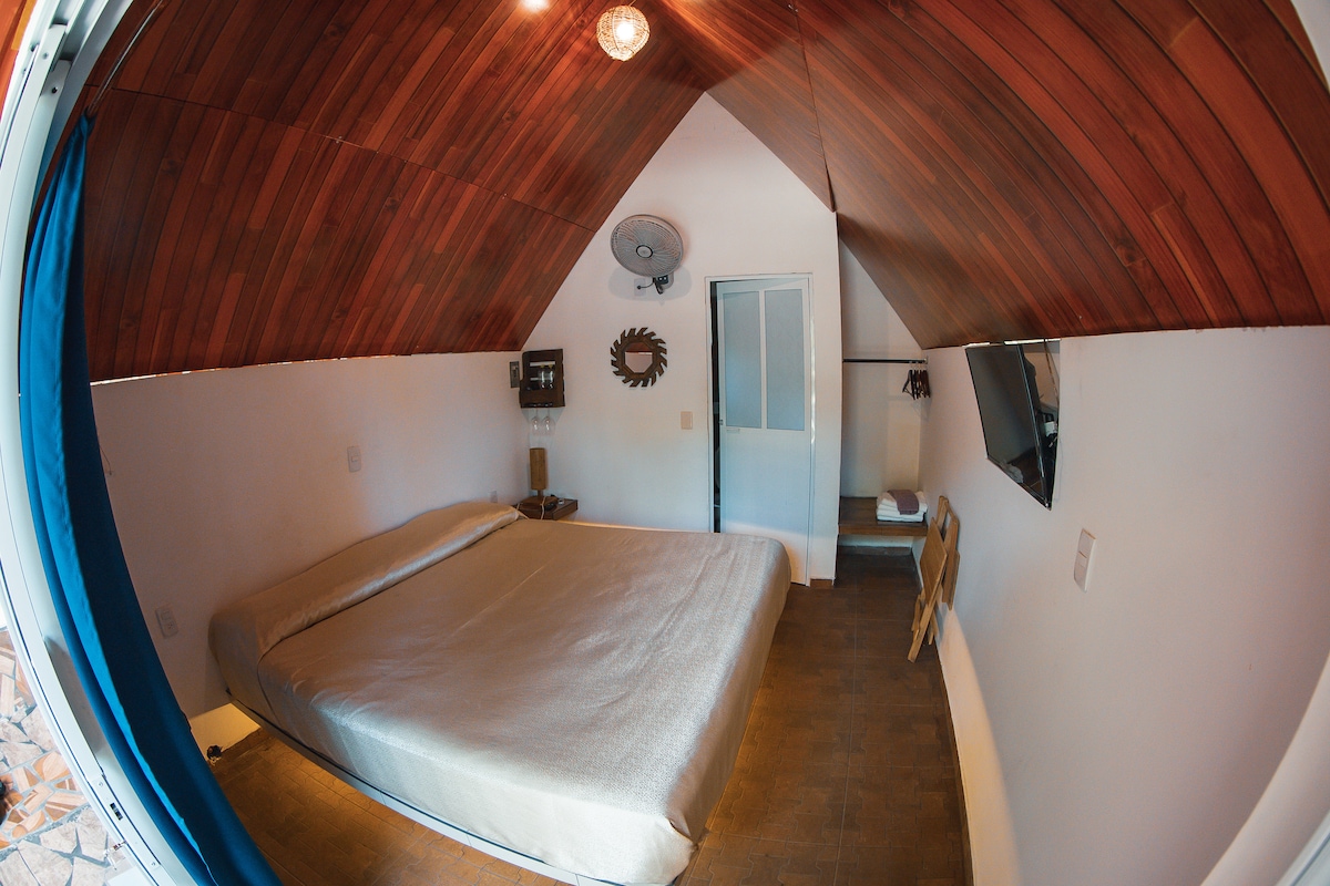 A spacious room features a king-size bed covered with a light-colored sheet, set against a wooden ceiling with natural tones. A television is mounted on the wall, while a small chair and mirror add functionality. The room is lit by both natural and artificial light.