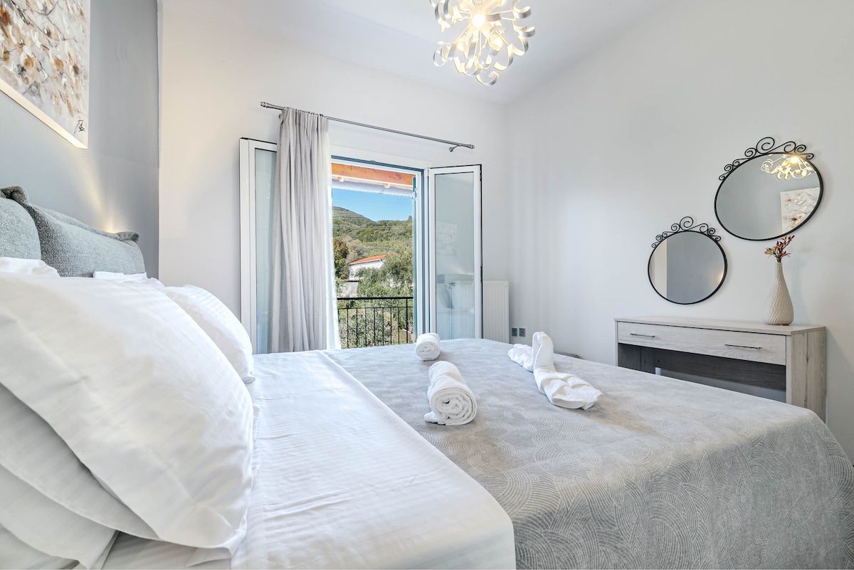 A well-appointed bedroom features a spacious bed adorned with neatly arranged towels. Natural light floods the room through double doors that open to a balcony with scenic views. A stylish mirror and a wooden dresser add to the room's aesthetic.