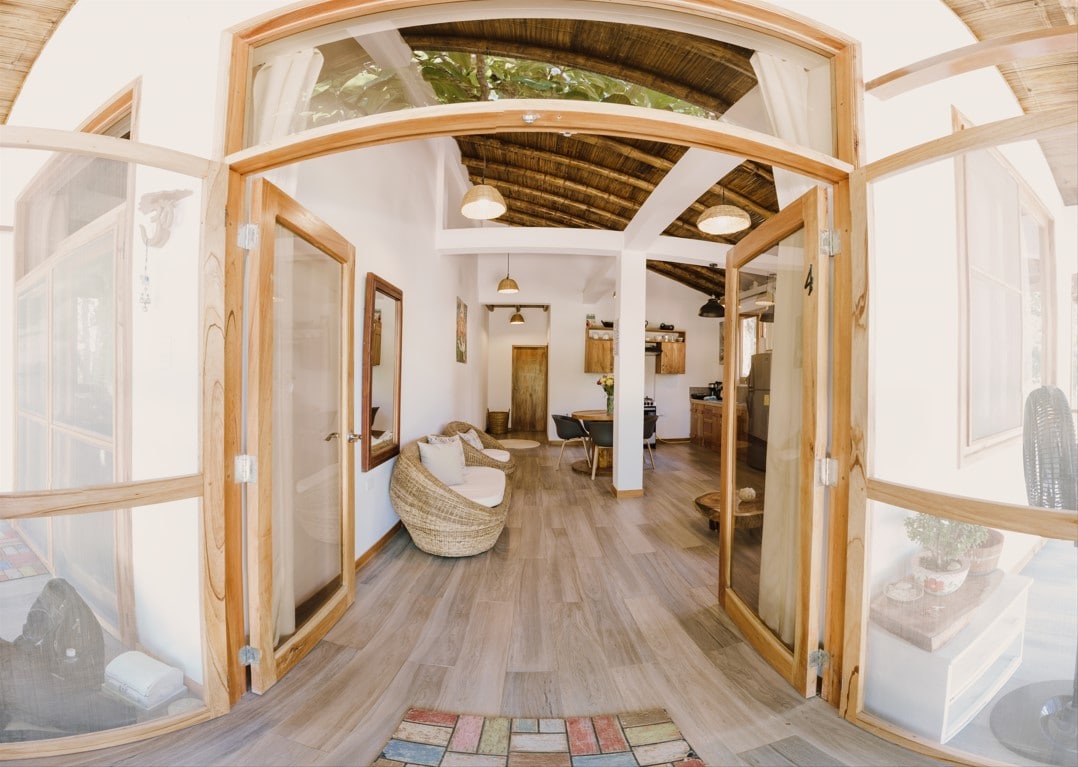 An inviting entryway features large wooden doors opening into a bright, airy living space. Natural light floods the room, highlighting a cozy seating area and rustic wooden beams overhead. The open layout seamlessly connects to a kitchen area in the background.
