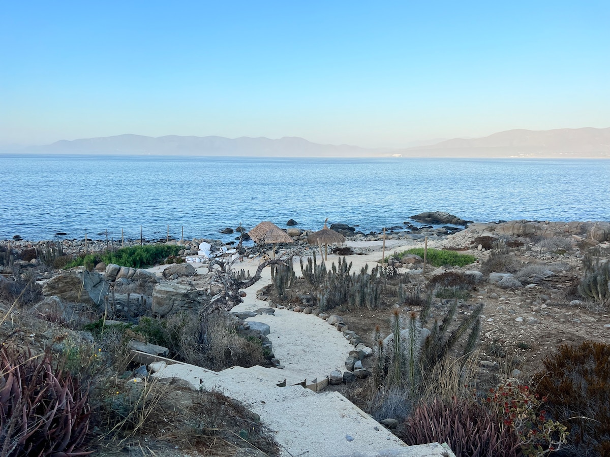 A sandy pathway meanders through coastal vegetation leading to the shimmering sea. Rocky formations and cacti are visible on either side, while gently rolling hills can be seen in the distance, all under a clear blue sky.