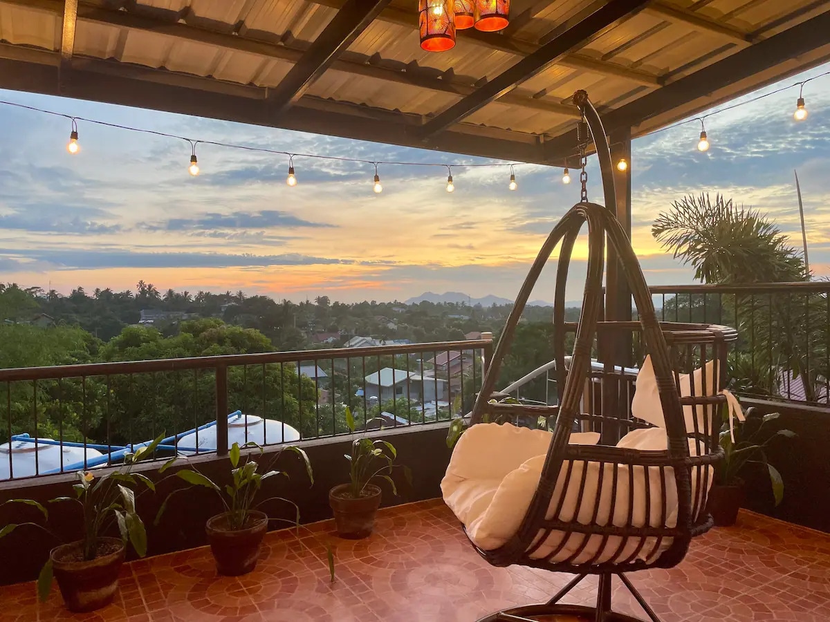A hanging chair is positioned on a veranda, offering a view of the horizon at sunset. String lights illuminate the area, with potted plants placed along the railing. A tranquil atmosphere is created by the surrounding landscape and distant mountains.
