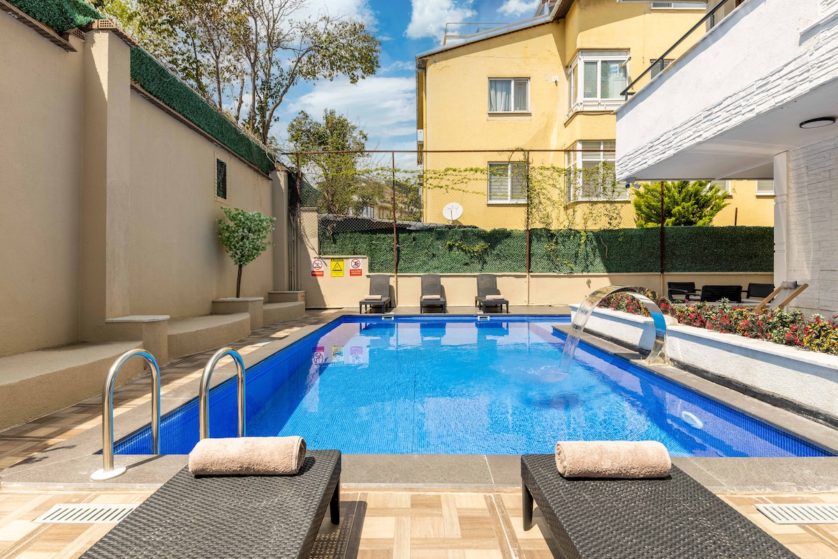 A refreshing outdoor pool is framed by a spacious deck. Two lounge chairs are positioned poolside, with towels neatly rolled placed on each. Lush greenery and flower beds enhance the setting, while the backdrop features a softly colored building under a clear blue sky.