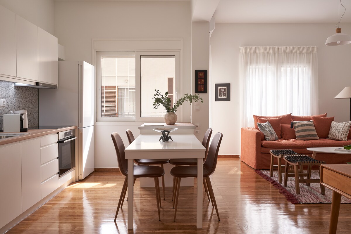 A modern kitchen area is displayed with sleek cabinetry and appliances, featuring a spacious dining table surrounded by wooden chairs. Natural light enters through a large window, illuminating the space. A comfortable seating arrangement, with warm-toned cushions, is visible in the living area beyond.