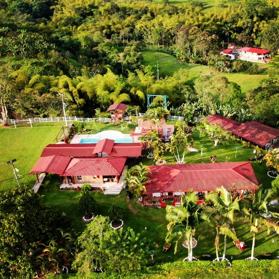 An aerial view captures a vibrant property featuring a central pool surrounded by lush green grass and tropical plants. The buildings, topped with red roofs, are positioned among expansive landscaped grounds, providing a serene atmosphere amid the surrounding natural landscape.