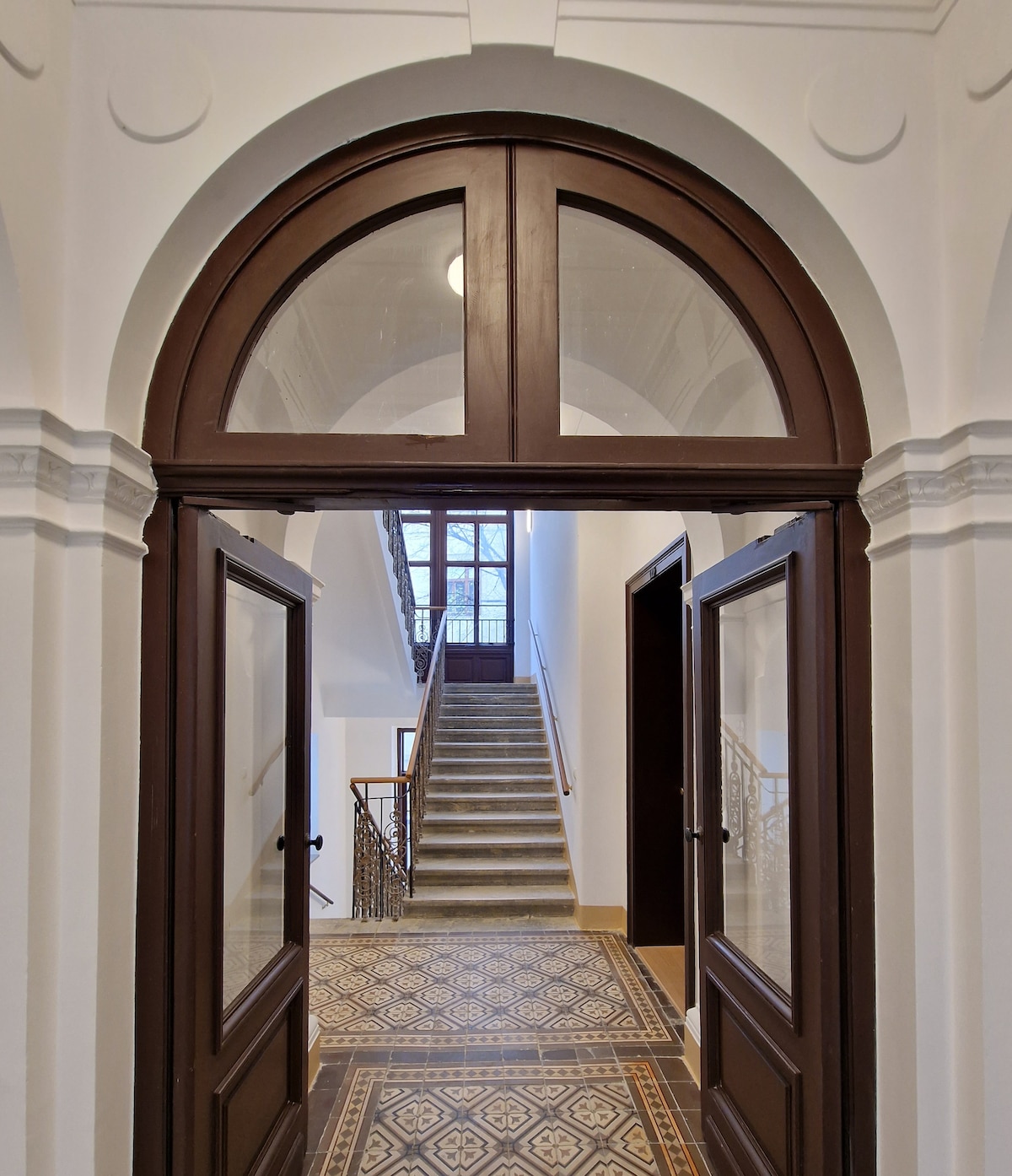 An elegantly arched wooden doorway opens into a well-lit hallway, showcasing a sweeping staircase with a decorative wrought-iron railing. A patterned tiled floor adds character to the space, while tall windows at the top of the stairs allow natural light to filter in.