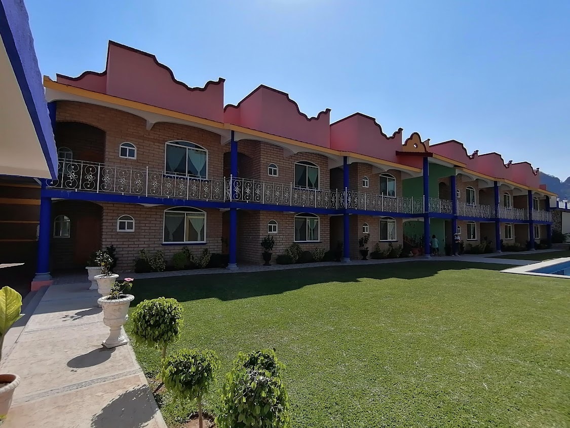 A row of colorful bungalows is displayed, featuring balconies with ornate railings. Flanked by well-maintained garden areas, the bungalows are situated alongside a large pool, under a clear blue sky. Each unit is designed with large windows for natural light.