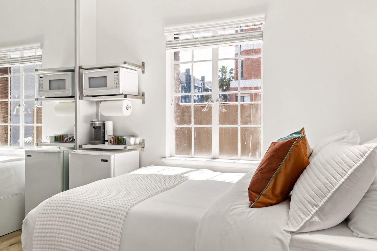 A bright and airy studio features a spacious bed with neatly arranged pillows. Natural light filters through large windows, illuminating the room. A mini fridge and microwave are positioned nearby, enhancing the smart design of the compact space.