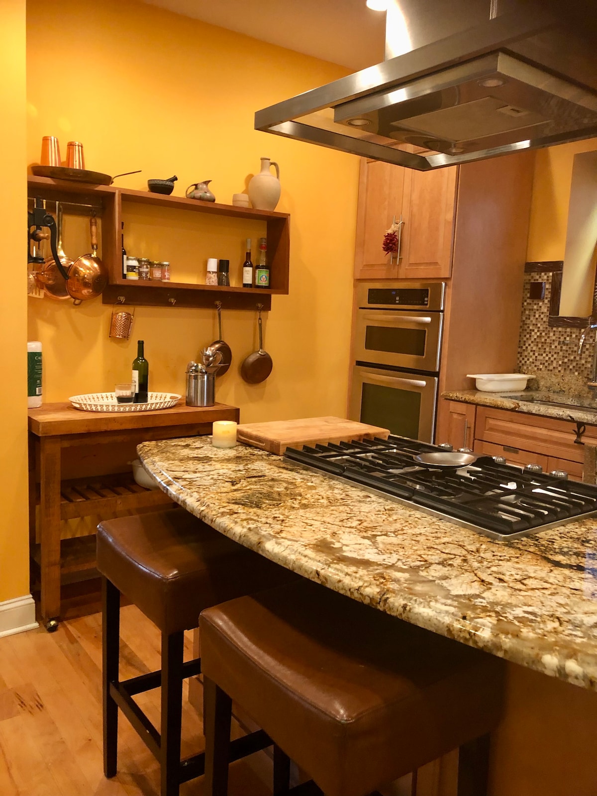 A well-equipped kitchen features a striking granite countertop with two bar stools. Warm yellow walls complement wooden cabinetry and a hanging shelf displaying cooking utensils. Premium appliances, including a double oven and a stovetop under a sleek hood, enhance the culinary space.