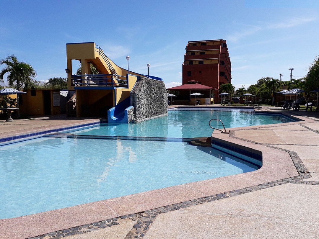 A swimming pool is set against a clear blue sky, featuring a slide on one side and surrounded by lounge areas and umbrellas. A multi-story building stands in the background, providing a contrast to the bright water and outdoor space.