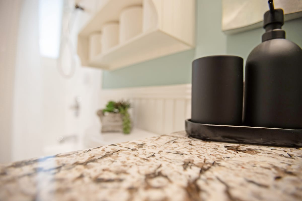 A close-up image of a granite countertop showcases a smooth, textured surface with natural patterns. Black cylindrical containers are positioned on a tray, alongside a small decorative plant in a woven basket. Soft light illuminates the space, highlighting the countertops and bathroom fixtures in the background.