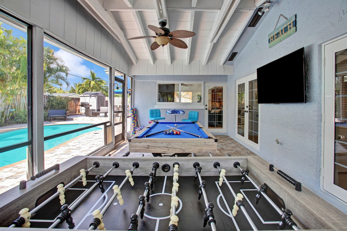 A bright Florida room features a foosball table and a pool table, surrounded by large glass windows that provide views of the outdoor pool area. The space is well-lit, with a ceiling fan overhead and comfortable seating visible in the background.