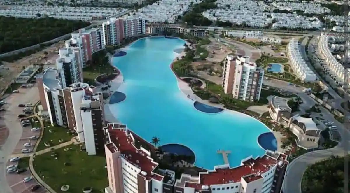 Aerial view of a sprawling pool area, featuring a winding shape that meanders through the landscape. Surrounding buildings are designed with a modern architectural style, complemented by lush greenery and pathways. The clear water reflects the sunny sky, creating a serene atmosphere.