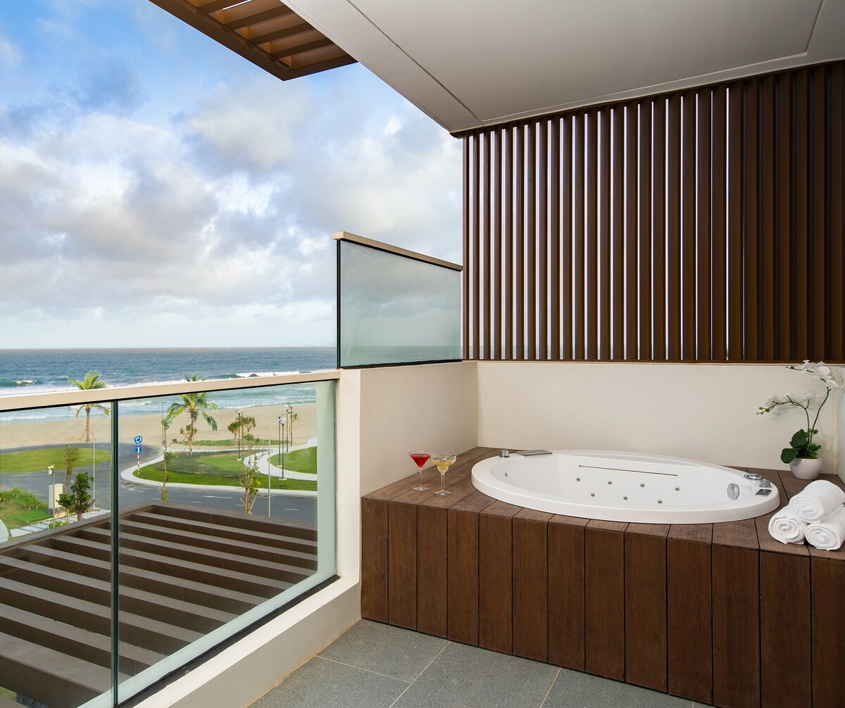 A private outdoor area features a hot tub, surrounded by sleek wooden paneling and an unobstructed view of the ocean. A large glass railing provides an open feel, while a small table holds two drinks. Fresh towels and plants add a touch of elegance.
