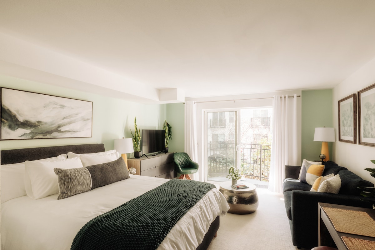 A spacious room features a large bed with a dark green throw, accompanied by a plush sofa and decorative armchair. Natural light floods in through the sliding glass door, revealing a balcony. Simple artwork adorns the walls, enhancing the modern decor.