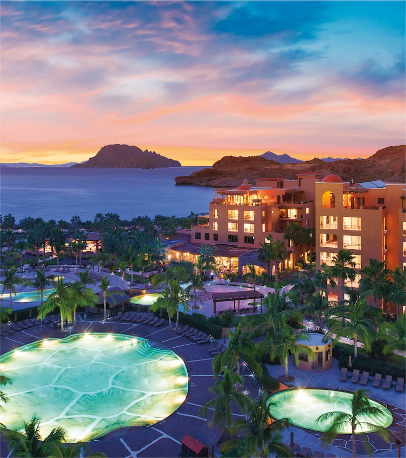 The resort complex is surrounded by lush palm trees and features expansive outdoor pools. A setting sun casts soft colors over the scene, reflecting off the calm waters of the sea. The distant mountains and islands add depth to the panoramic view.