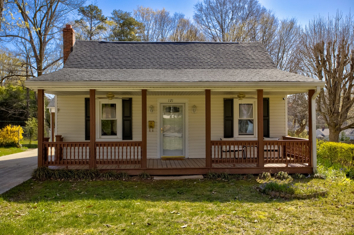 The charming exterior of the cottage features a welcoming front porch with wooden railings and ample seating. The cozy facade is accented by well-maintained greenery and large windows dressed in dark curtains, inviting natural light into the home.
