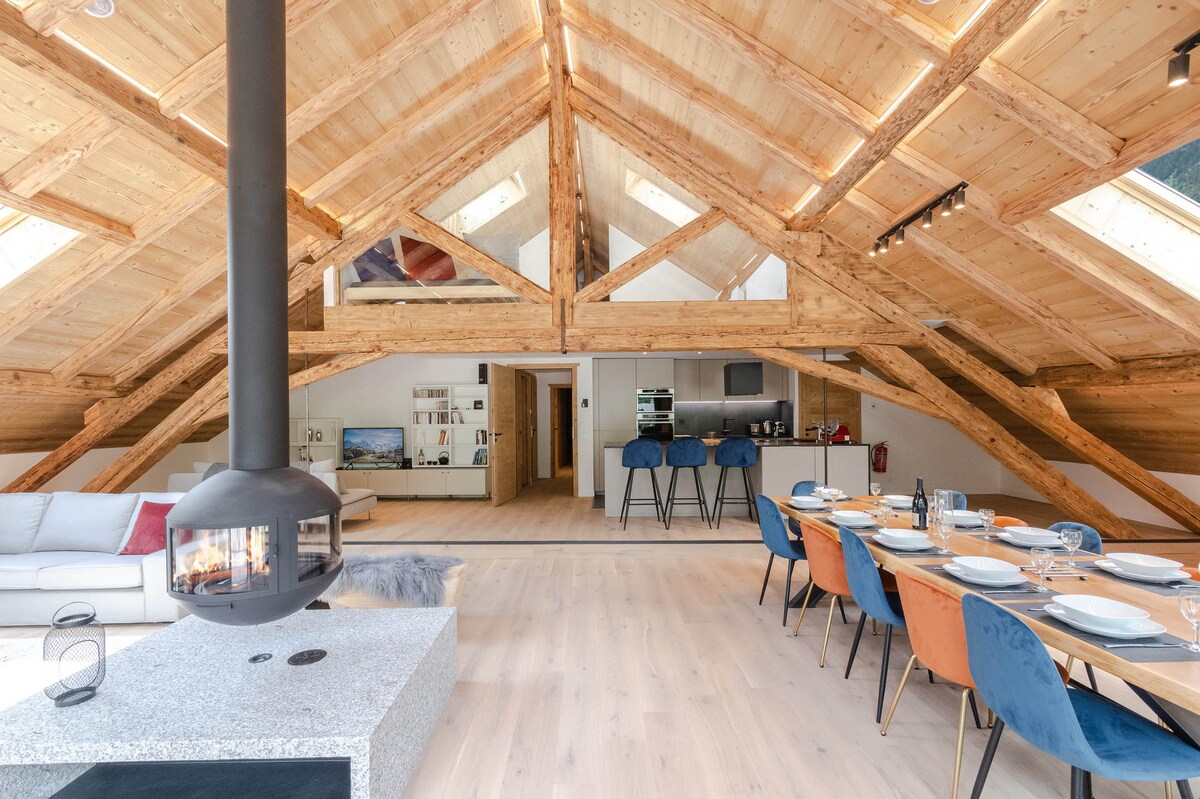 An open-concept living space features vaulted wooden ceilings and ample natural light from skylights. A modern dining table is set for several guests, surrounded by colorful chairs. A stylish fireplace stands centrally, complementing the cozy atmosphere of the loft's design.