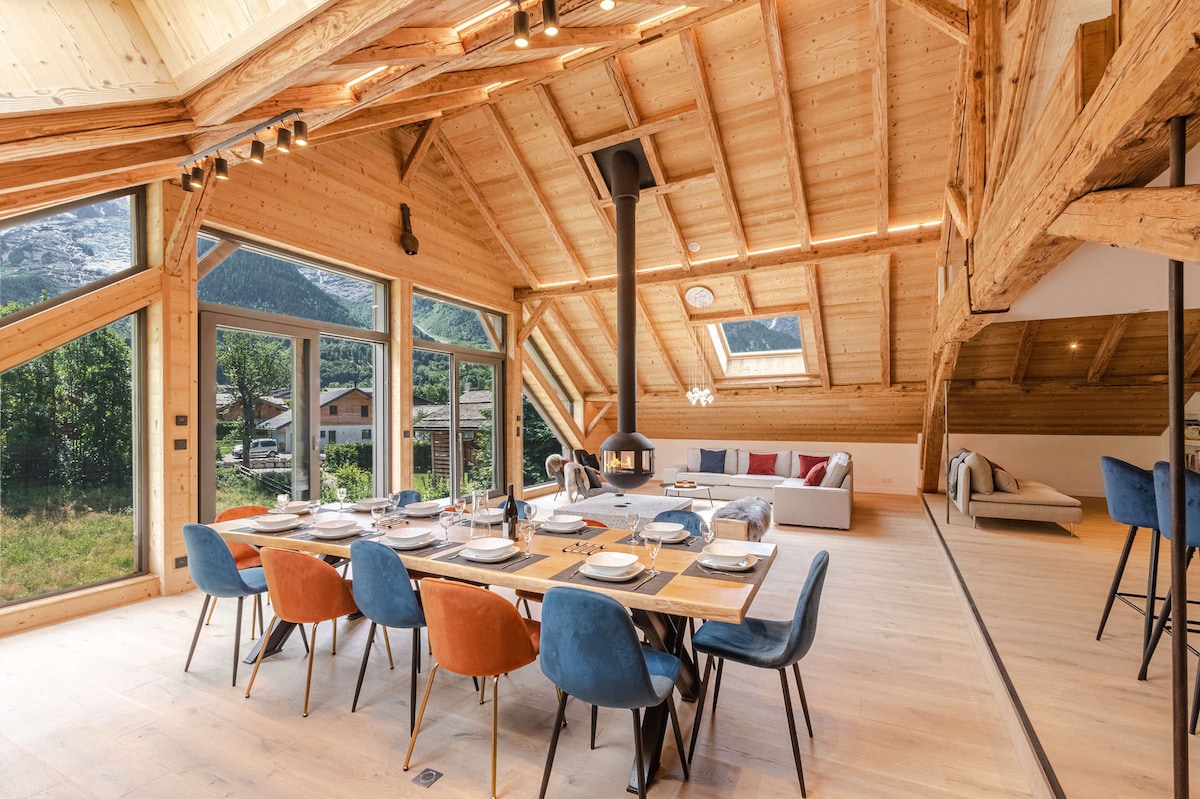 A spacious dining area features a large wooden table surrounded by multi-colored chairs. Large windows provide views of the outdoor landscape. A modern fireplace is centrally located, enhancing the cozy atmosphere. Natural light fills the room through the sloped wooden ceiling.