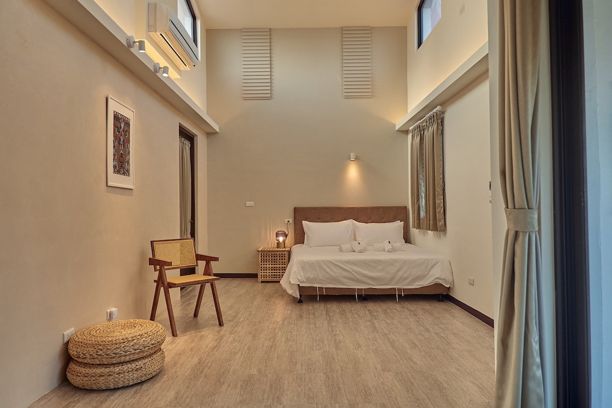 A spacious room features a large bed with soft linens against a neutral backdrop. A wooden chair and a round woven mat add a touch of natural texture. Natural light enters through windows, creating a serene ambiance throughout the space.