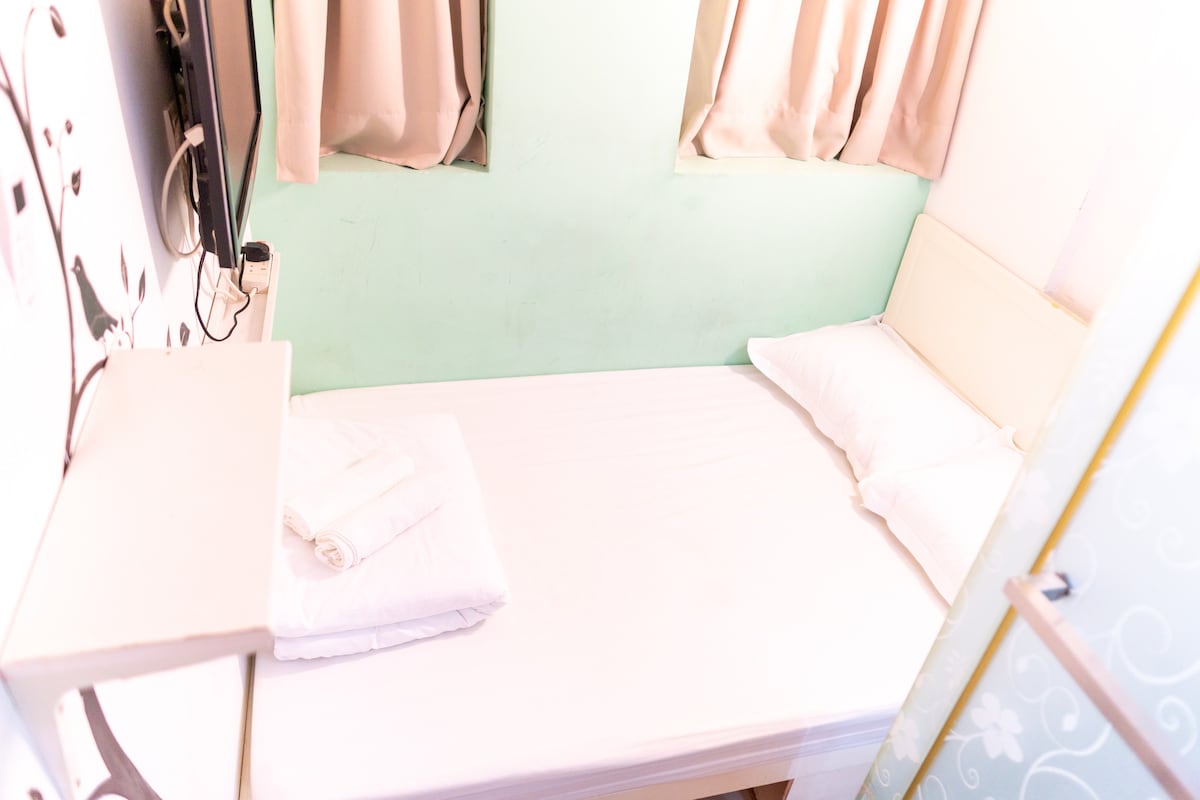 A compact sleeping area features a white bed with two pillows and fresh linens. Two windows with light curtains allow natural light to fill the space. The walls are painted a soft green, and a small shelf is attached to one side.
