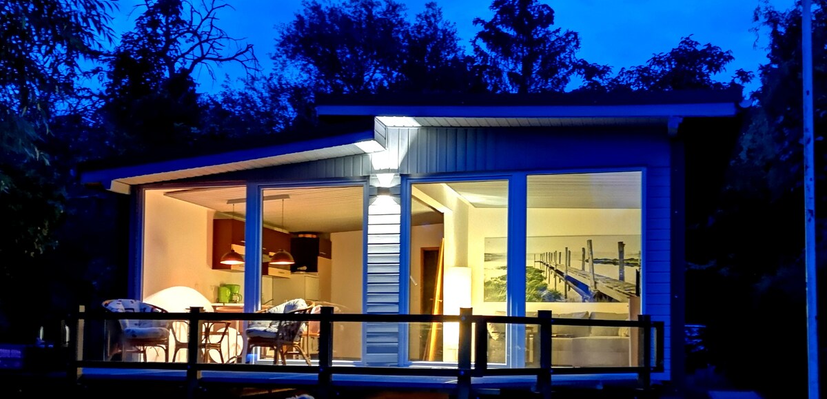 The exterior of a modern boathouse is presented at twilight, featuring large windows that illuminate the interior space. The outdoor deck showcases comfortable seating, enhancing the connection with the surrounding natural landscape.