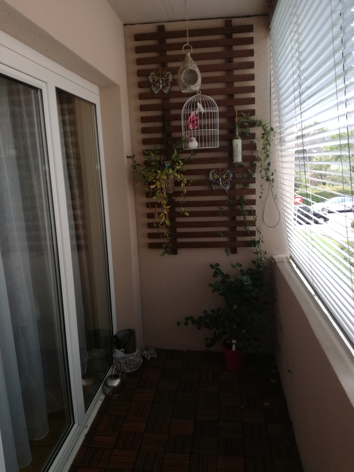 A cozy balcony is presented with a wooden panel decorated with hanging plants and a birdcage. Sunlight streams through the horizontal blinds, illuminating the potted greenery at the base. A tiled floor adds texture to the space, enhancing the inviting atmosphere.