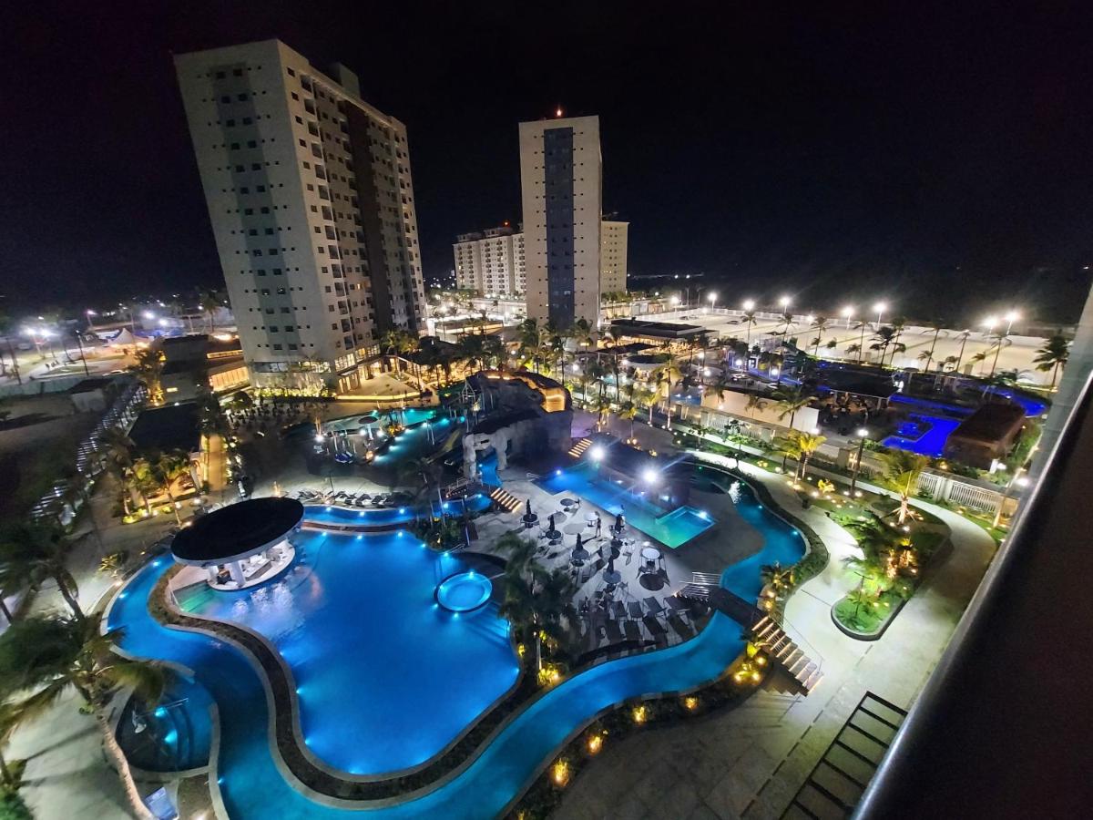 A panoramic view showcases the beautifully illuminated resort area at night. A winding pool is surrounded by lush greenery, with lounge chairs positioned nearby. The surrounding buildings stand tall against the dark sky, reflecting the vibrant lighting that enhances the social atmosphere.