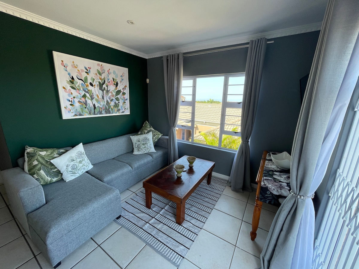 The lounge area features a spacious gray sofa adorned with decorative cushions, facing a wooden coffee table. A piece of colorful artwork is displayed on the green accent wall, while soft gray curtains frame the window, allowing natural light to brighten the room.