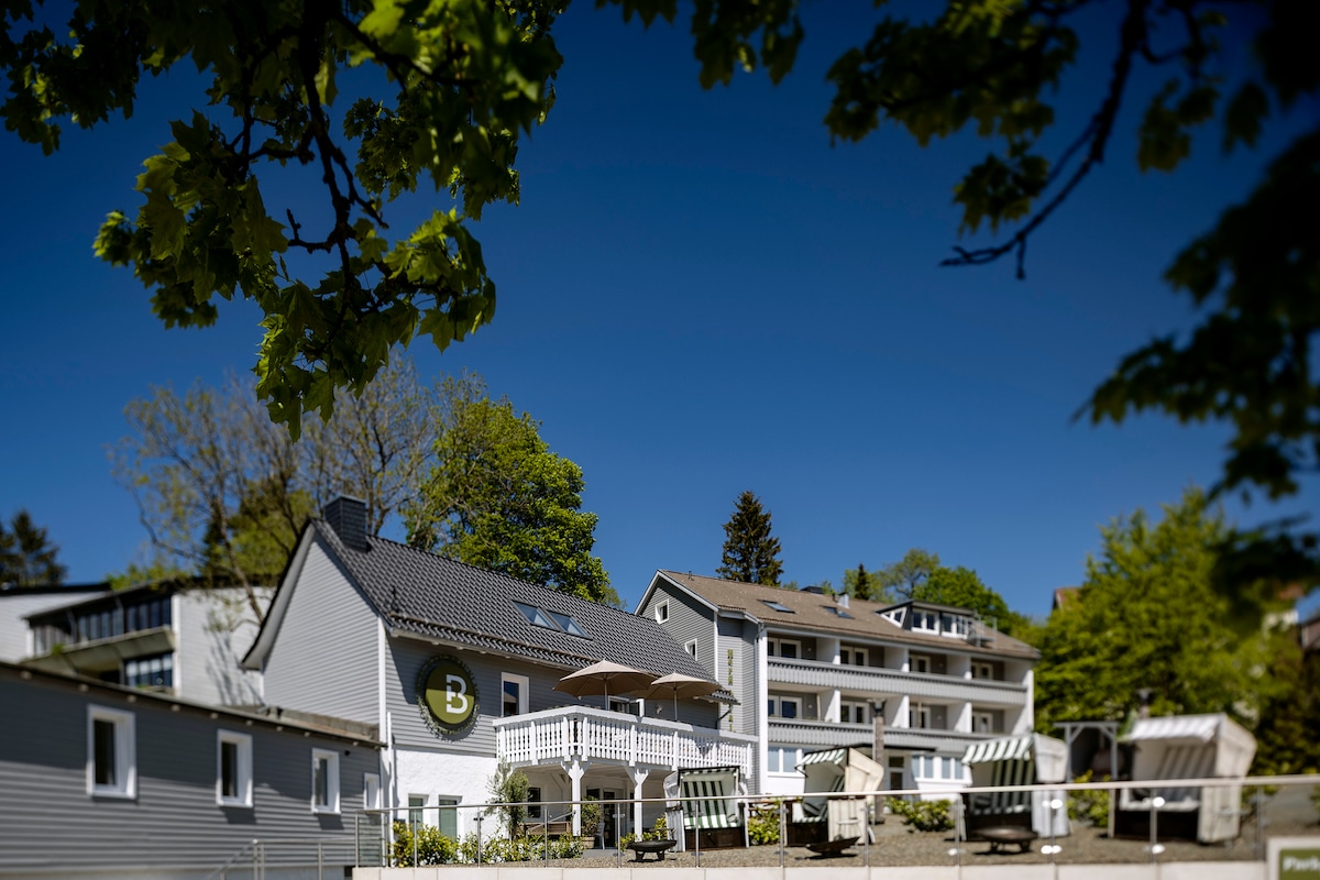 A collection of buildings includes a charming two-story house featuring a balcony and a large letter 'B' on its facade. Surrounding trees provide shade, and a clear blue sky creates a bright atmosphere, enhancing the inviting setting.