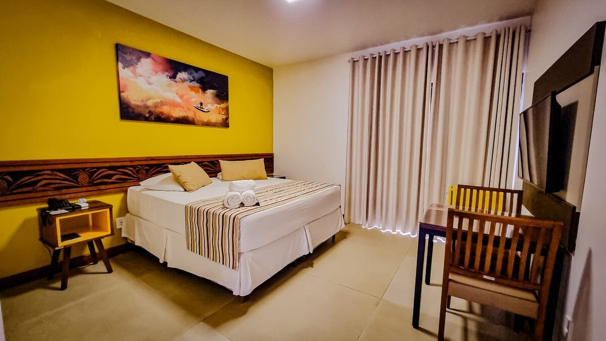 A comfortable guest room features a spacious bed with neutral linens and decorative pillows. An artwork piece above the bed showcases soft hues. A small wooden desk and chair are positioned near the window, which is dressed with light-colored curtains, allowing natural light to filter in.