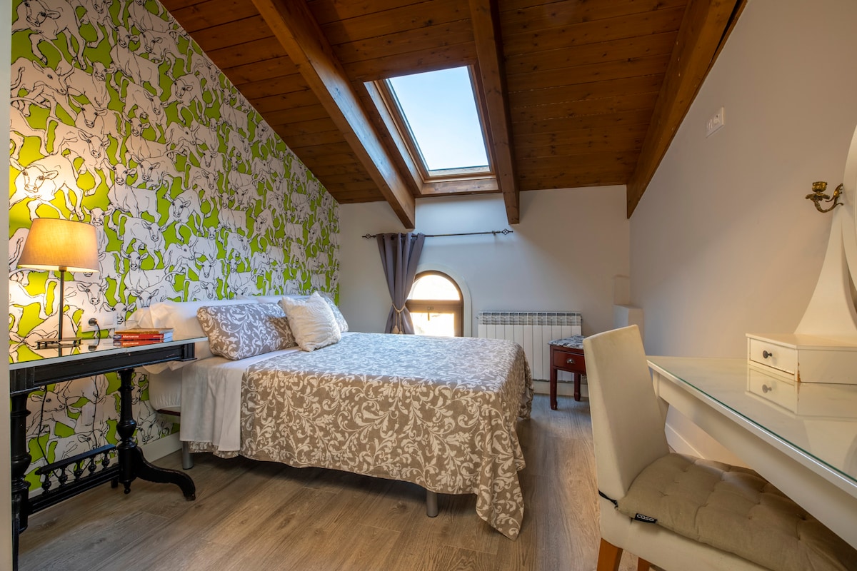 A private attic room features wooden beams and a sloped ceiling with skylights allowing natural light. A spacious bed with a decorative blanket is positioned beside a vintage lamp and a small desk. A window with curtains provides views of the outside, enhancing the room's ambiance.