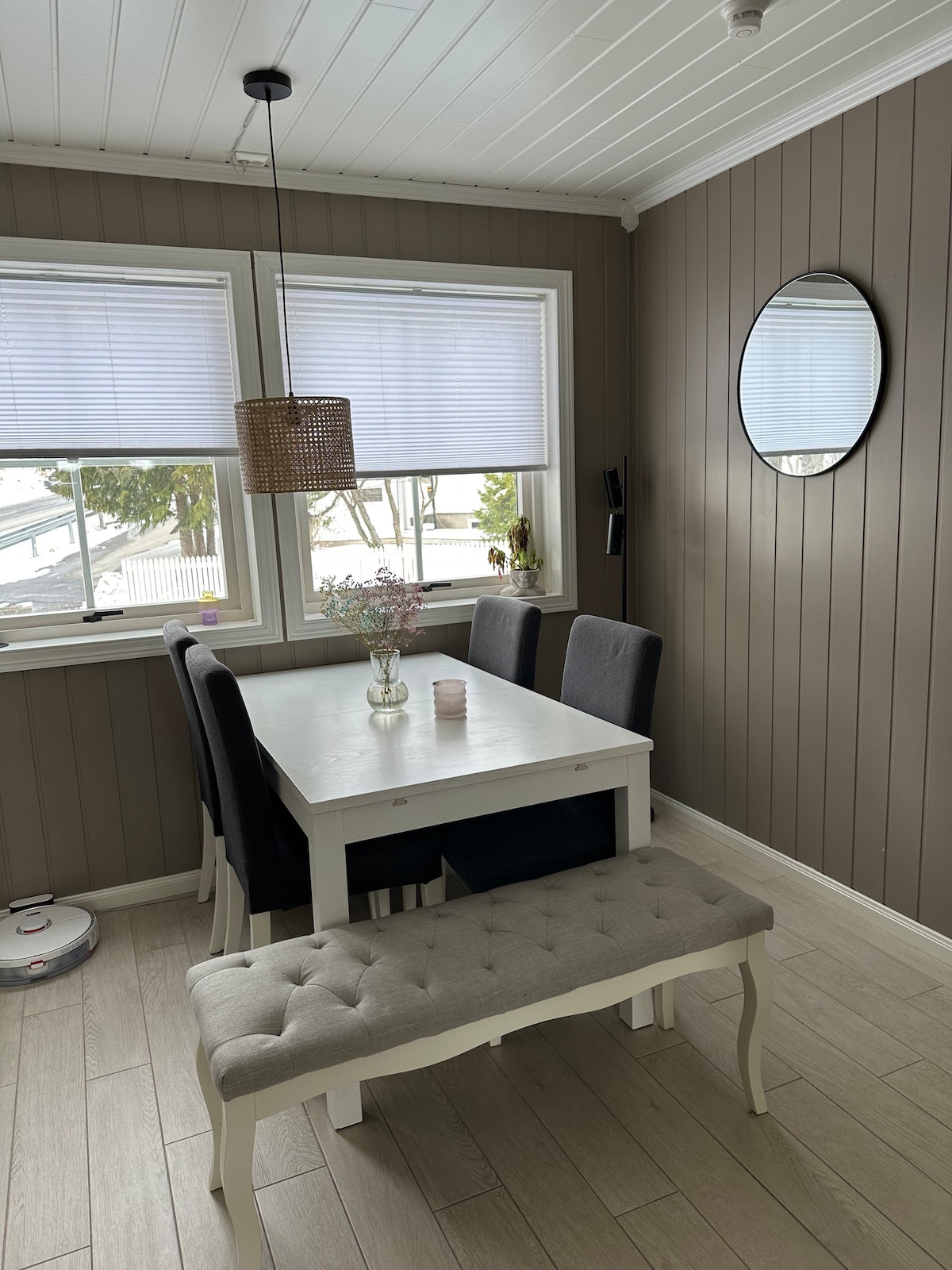 A dining area features a white rectangular table surrounded by four grey upholstered chairs. A bench adds extra seating, while two windows allow natural light to fill the room. A round mirror and a pendant light are mounted on the wall, enhancing the space.
