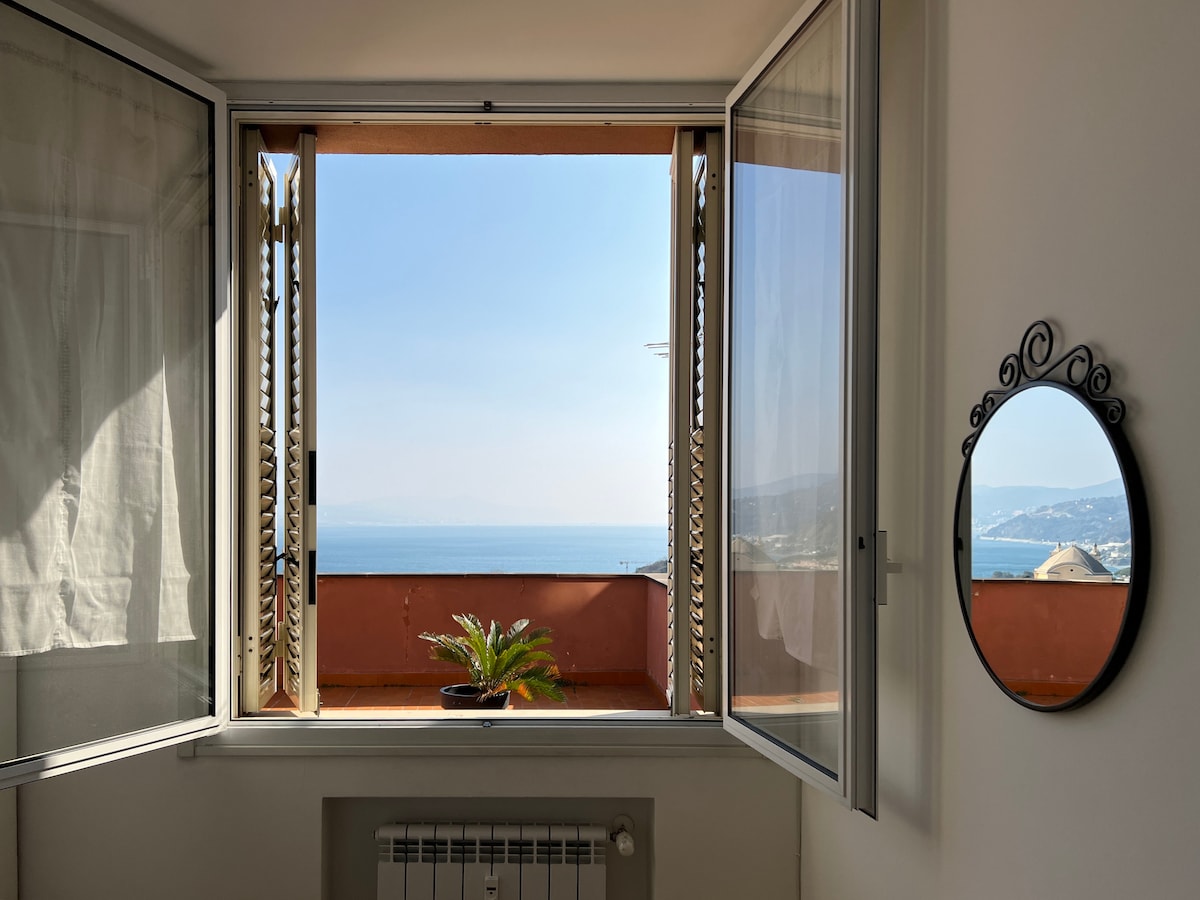 Two large windows open to reveal a panoramic view of the sea, with gentle waves meeting the horizon. A decorative mirror reflects the serene coastal scenery, while a potted plant adds a touch of green to the indoor space.