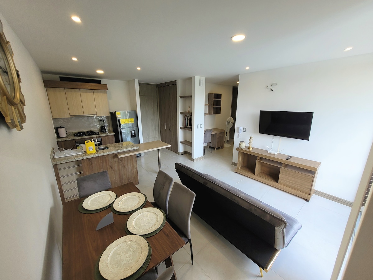 The open-plan living area is highlighted by a dining table set for four and a comfortable sofa facing a wall-mounted television. The kitchen features modern appliances and a breakfast bar, lending a spacious and functional feel to the room.