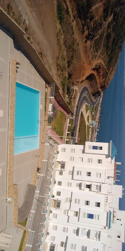 An outdoor swimming pool surrounded by a paved deck is visible, complemented by green landscaped areas. The building, featuring a modern architectural style, can be seen in the foreground, while a scenic roadway and hillside landscape are visible in the distance.