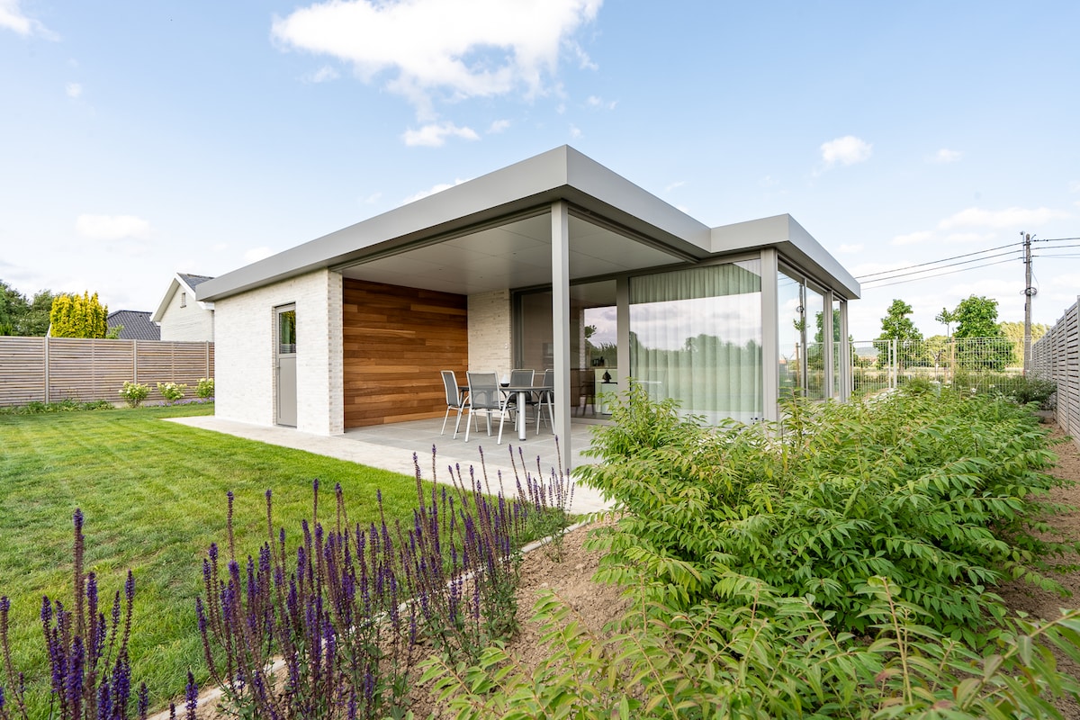 A modern bungalow is set amidst a landscaped garden, featuring a spacious terrace with outdoor furniture. Large glass doors provide a view of the airy interior, while surrounding greenery adds to the serene environment. The exterior combines light-colored brick and wooden accents.